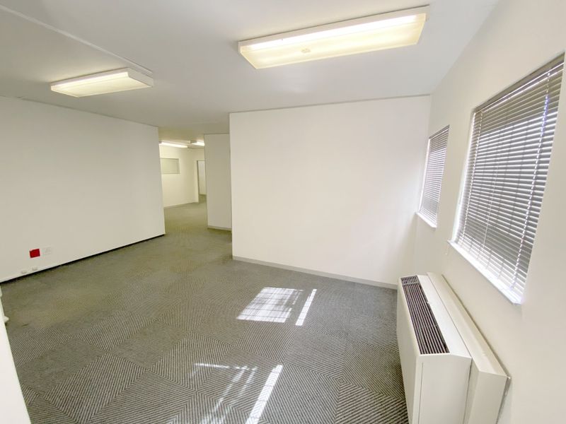 A clean, vacant commercial office space featuring carpeted flooring, built-in HVAC unit, and large windows with fitted venetian blinds.
