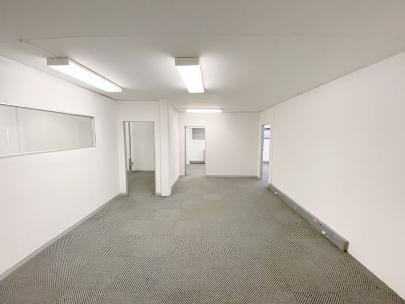 A clean, vacant commercial office space featuring neutral carpet tiles, bright overhead fluorescent lighting, and multiple access points to adjacent rooms.