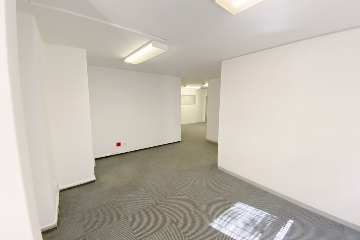 A vacant, open-plan commercial office space featuring neutral white walls, carpeted flooring, and overhead fluorescent lighting.