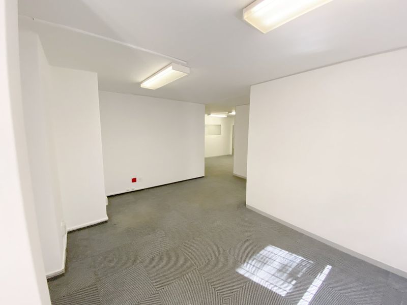 A vacant, open-plan commercial office space featuring neutral white walls, carpeted flooring, and overhead fluorescent lighting.
