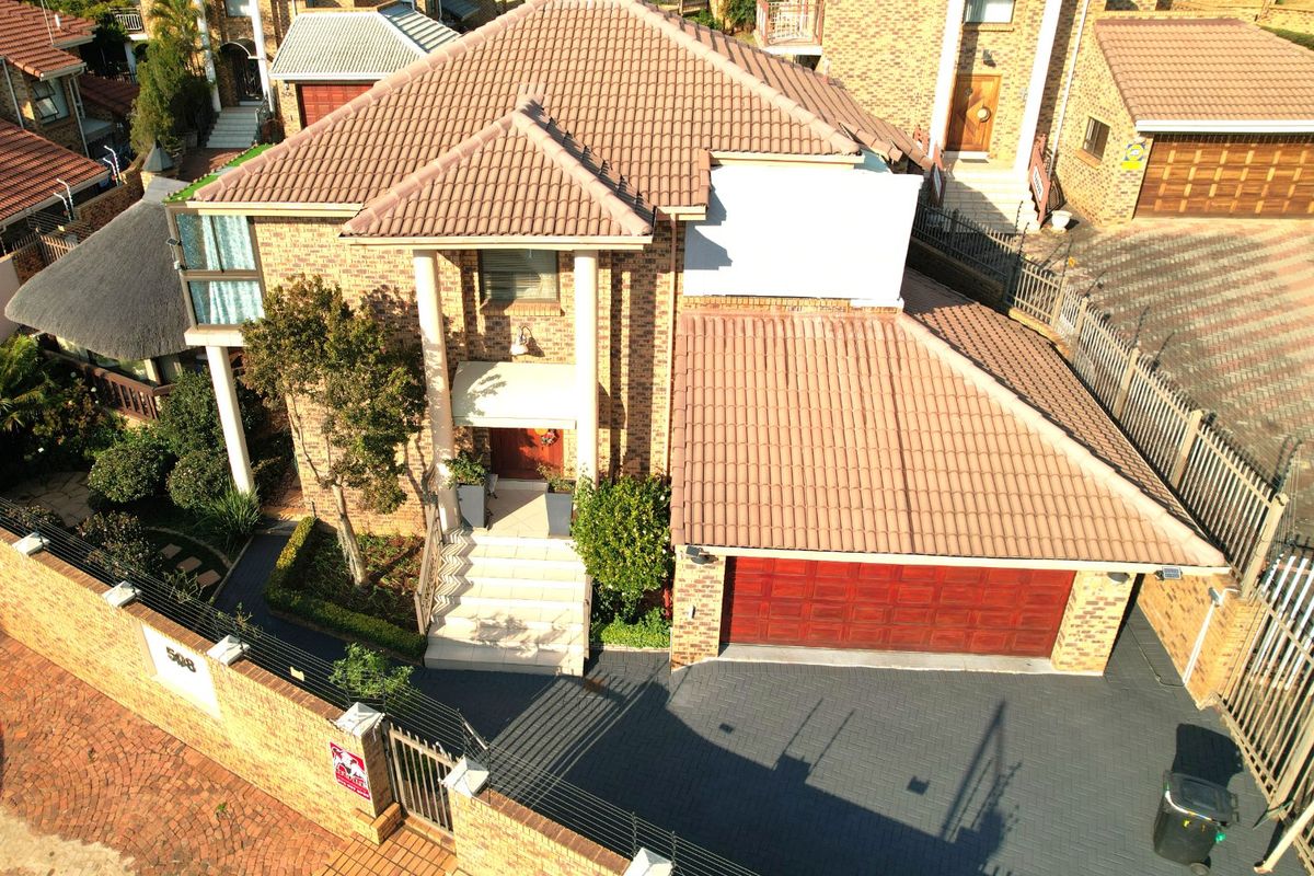 Aerial view of a double-story brick house featuring a tiled roof, paved driveway, double garage, and secure boundary wall with electric fencing.