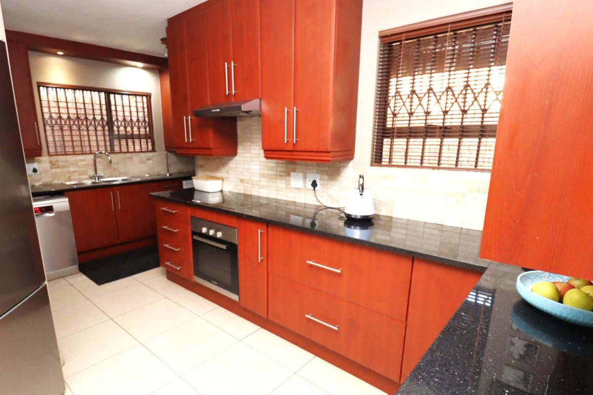 A functional kitchen featuring warm-toned wooden cabinetry, granite countertops, integrated appliances, and tiled flooring, complemented by natural light through window security bars.