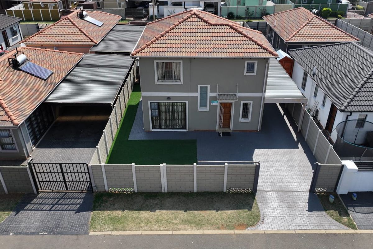 Aerial view of a two-story residential property featuring a paved driveway, carport, boundary wall, and artificial lawn.