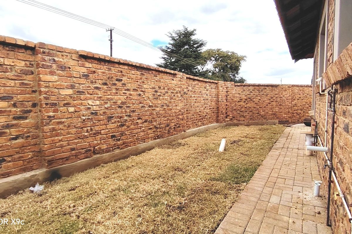 A rear exterior view of the townhouse featuring a face brick boundary wall, a paved walkway, and a small lawn area.