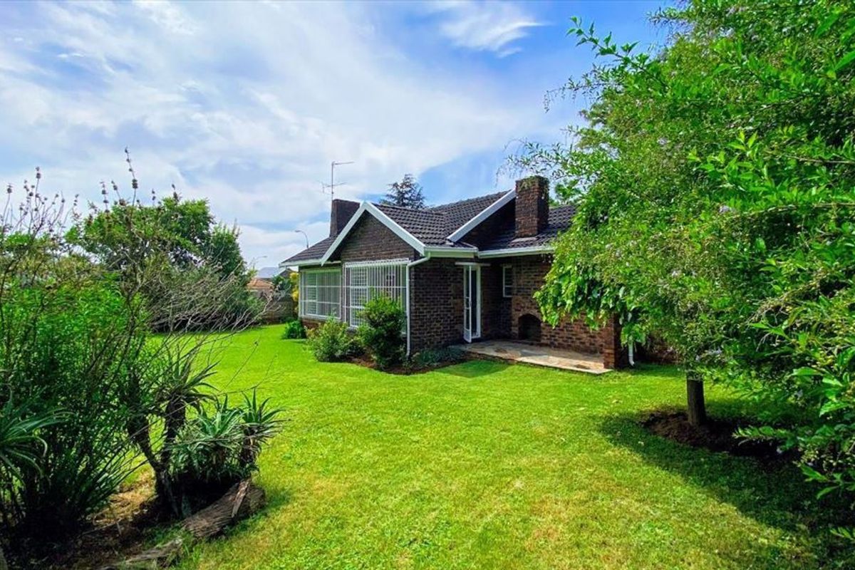 A view of the rear brick facade of the house, featuring a stone patio, tiled roof, and established garden surroundings.