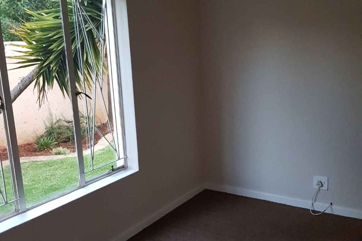 A vacant bedroom featuring neutral walls, carpeted flooring, and a large window with security bars overlooking the garden.