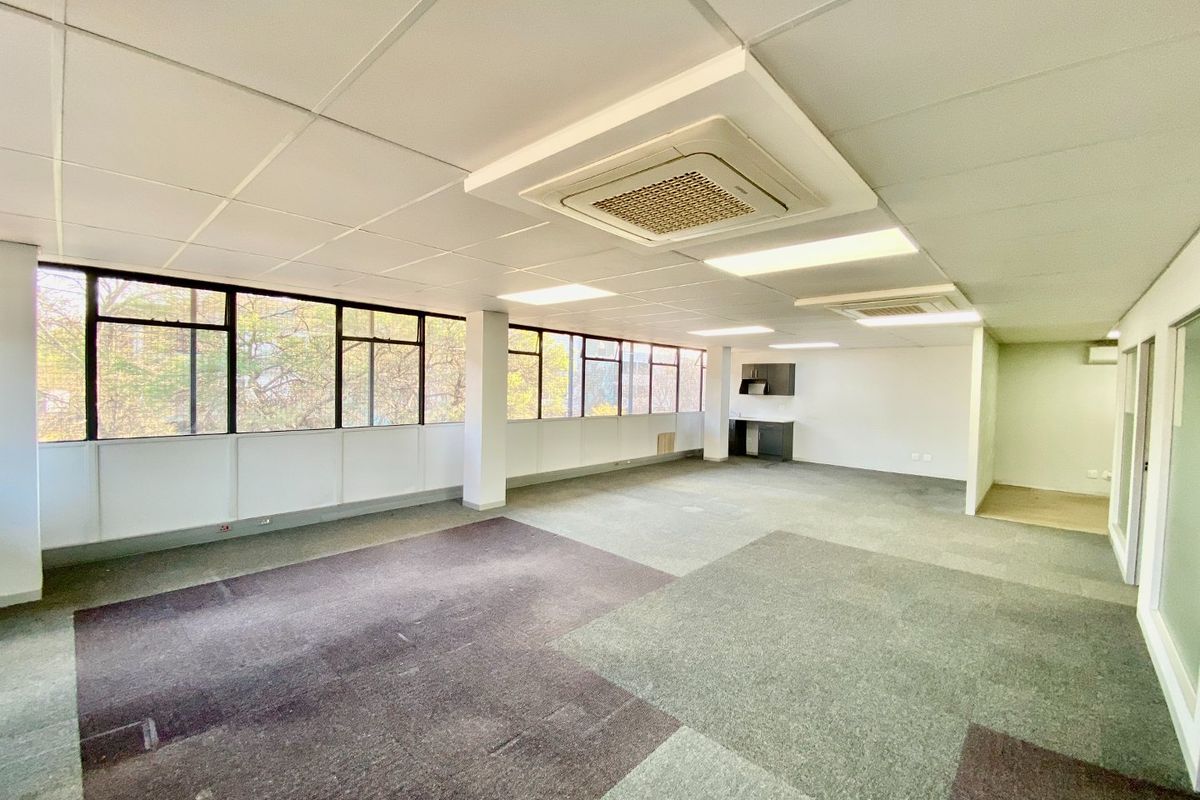 An open-plan commercial office space featuring large windows, suspended ceilings with integrated air conditioning, and a small kitchenette area.
