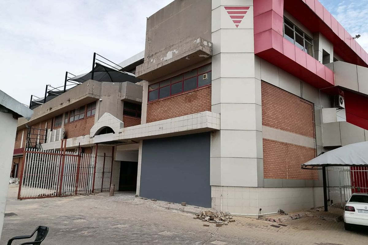 Rear exterior view of a commercial building featuring brick and metal cladding, security fencing, and a paved parking area.
