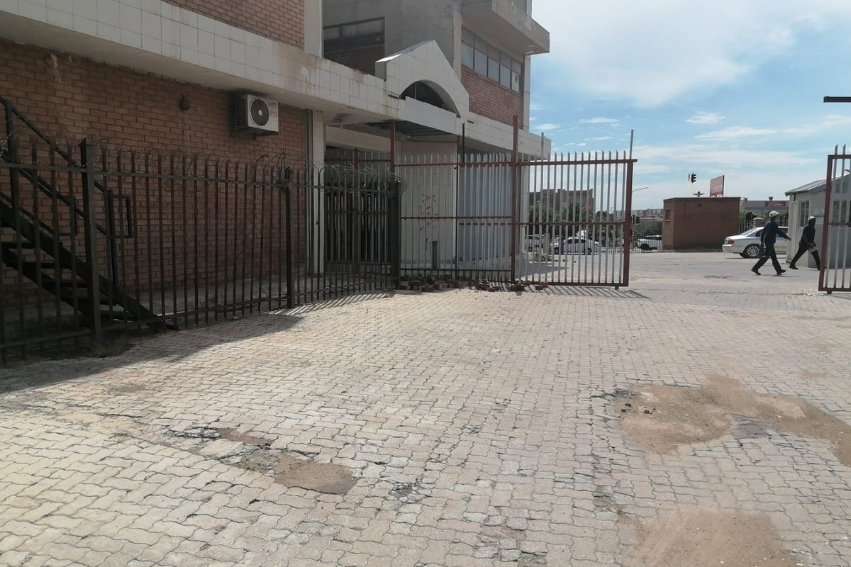 Front exterior view of a commercial building featuring brick facade, metal security fencing, and a paved driveway area showing signs of wear.