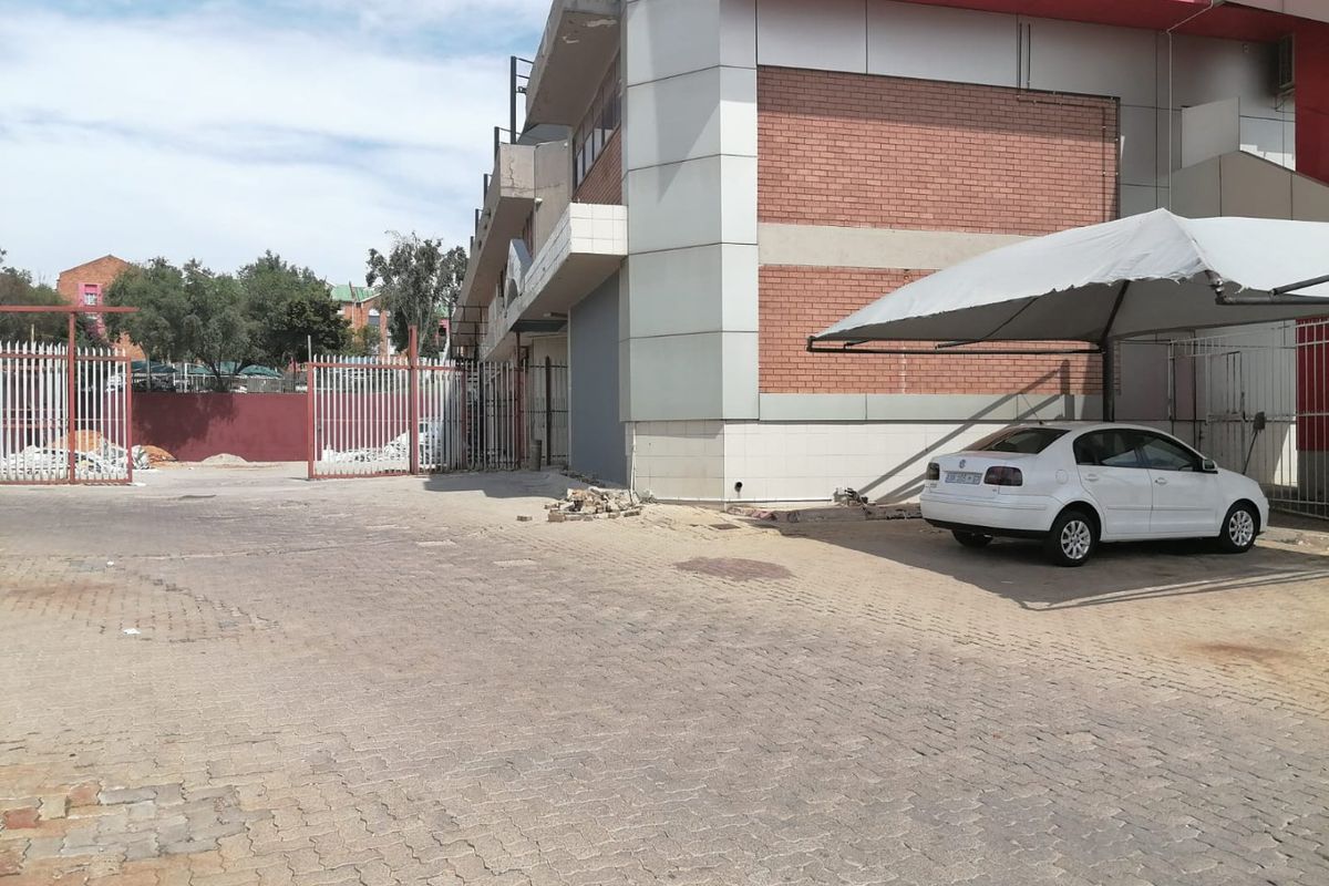 Rear exterior view of a commercial building featuring a paved parking area, metal security fencing, and a covered carport structure.