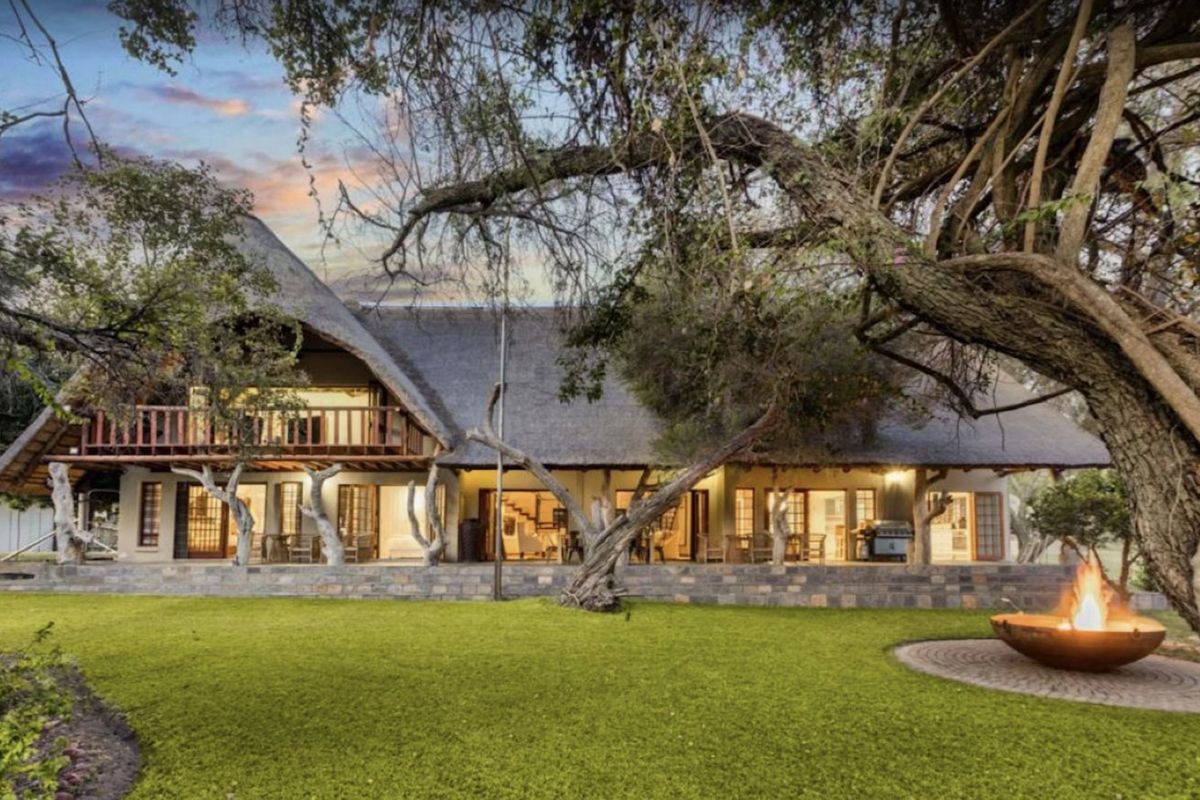 Rear view of a thatched-roof farm residence featuring a stone patio, upper-level balcony, and a landscaped garden with a built-in fire pit.