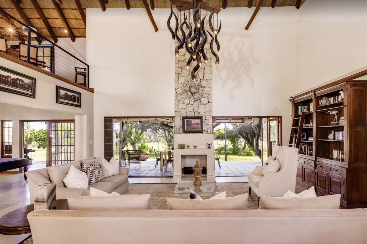 Spacious double-volume living room featuring a stone fireplace, exposed thatch ceiling, mezzanine level, and large folding glass doors opening to the garden.