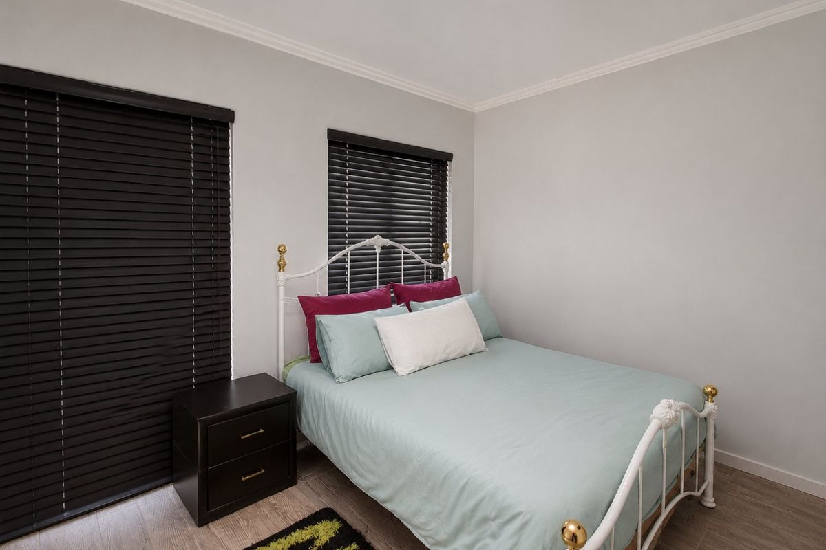 A well-maintained bedroom featuring light-toned laminate flooring, neutral wall finishes, and dark venetian blinds for privacy.