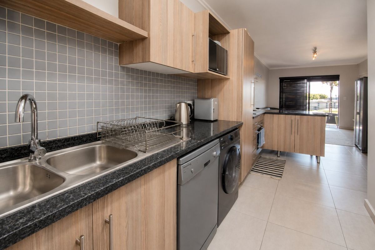 Modern kitchen featuring light wood-tone cabinetry, dark granite countertops, tiled backsplash, and integrated appliances, leading into an open-plan living area.