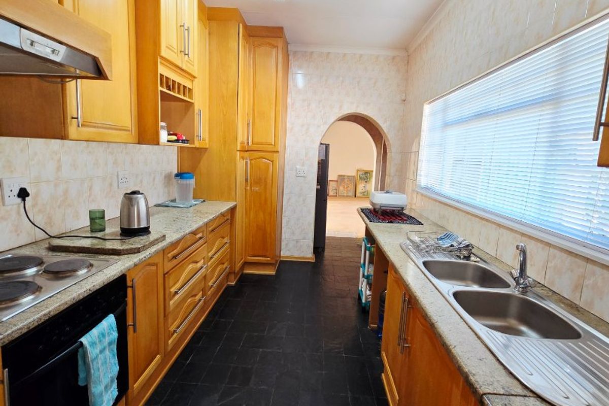 A functional galley-style kitchen featuring wooden cabinetry, granite countertops, a double sink, and slate flooring, leading through an archway to the living area.