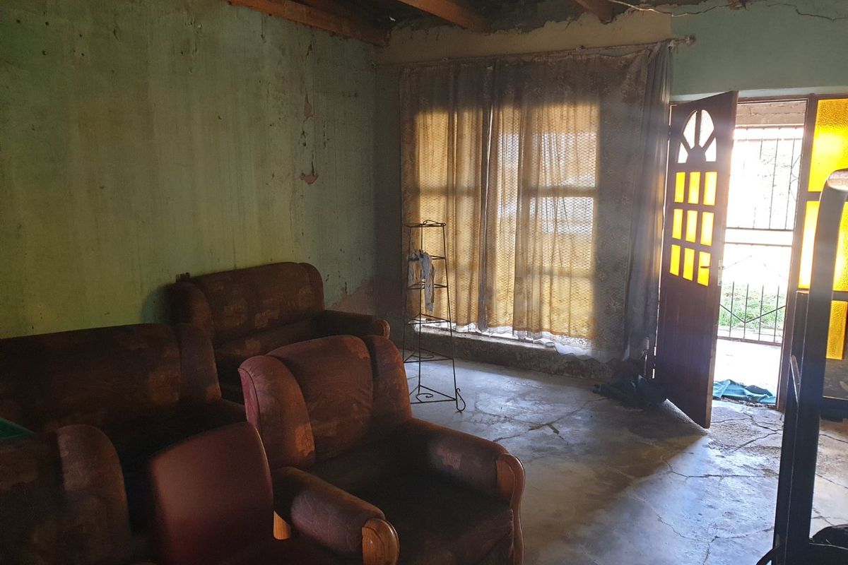 A living room space featuring exposed timber roof beams, concrete flooring, and a security gate, currently requiring significant renovation and cosmetic updates.