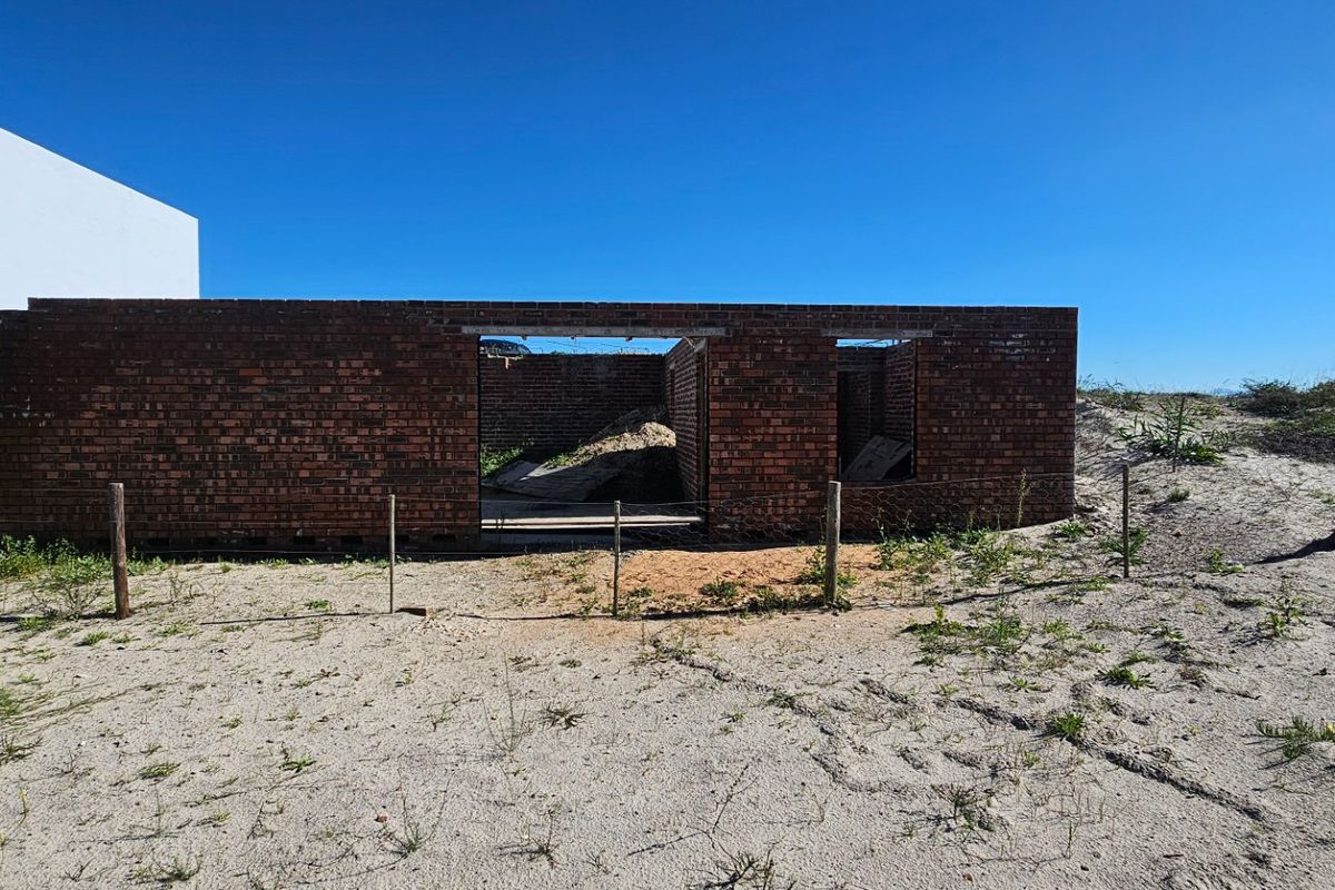 An exterior view of an incomplete brick structure under construction, showing exposed masonry walls and a sandy site environment.