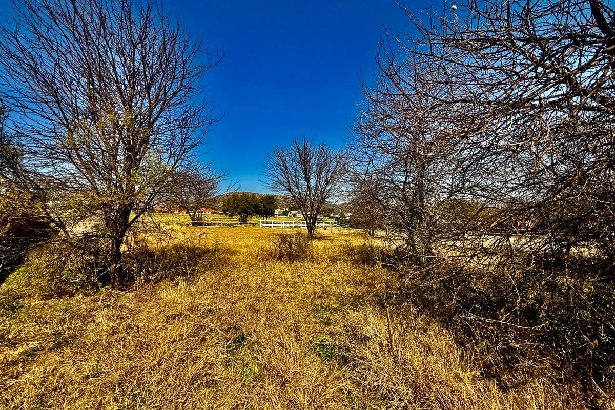 A view of a vacant residential plot featuring dry grass, mature trees, and a white post and rail fence in the distance.