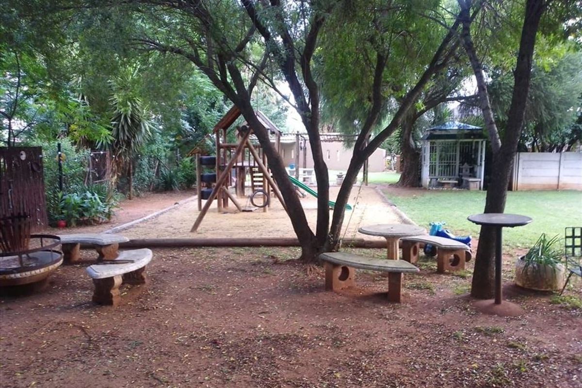 A spacious outdoor garden area featuring mature trees, a sand pit with a playground structure, concrete seating, and a small gazebo.