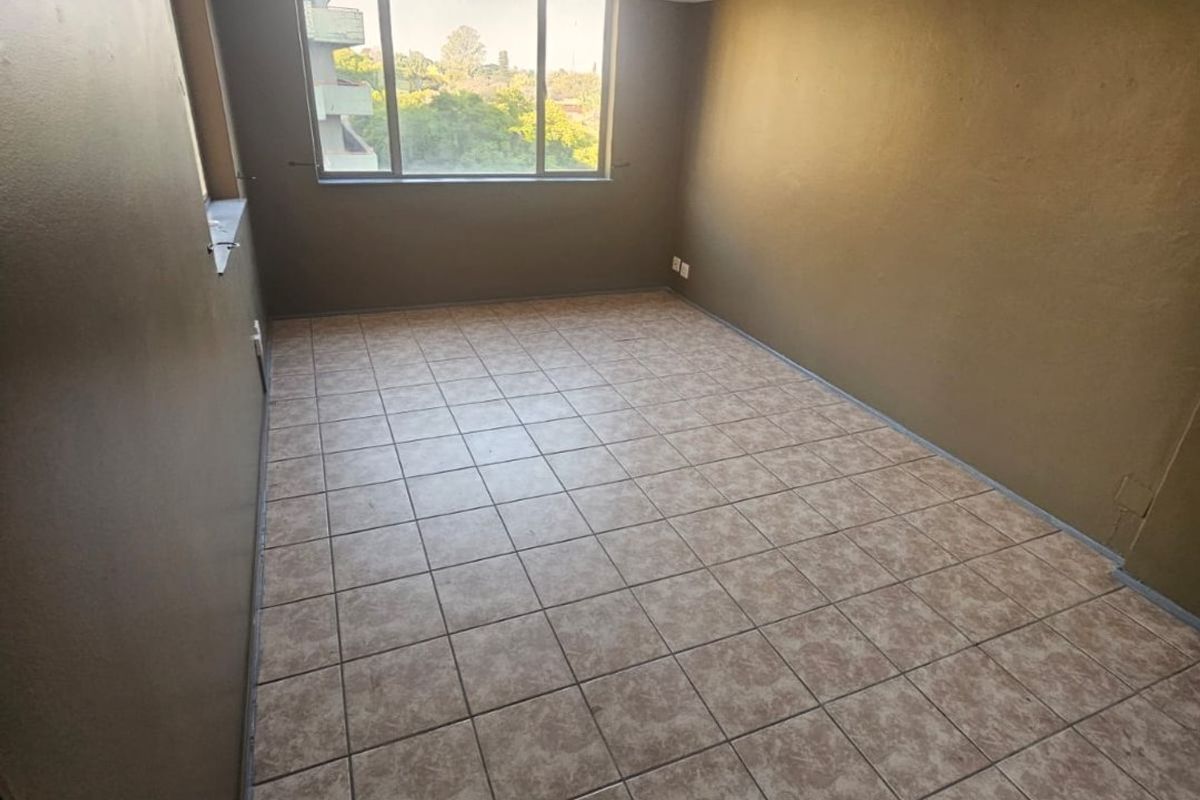 An empty, neutrally painted living area featuring tiled flooring, a textured ceiling, and a large window providing natural light and an exterior view.