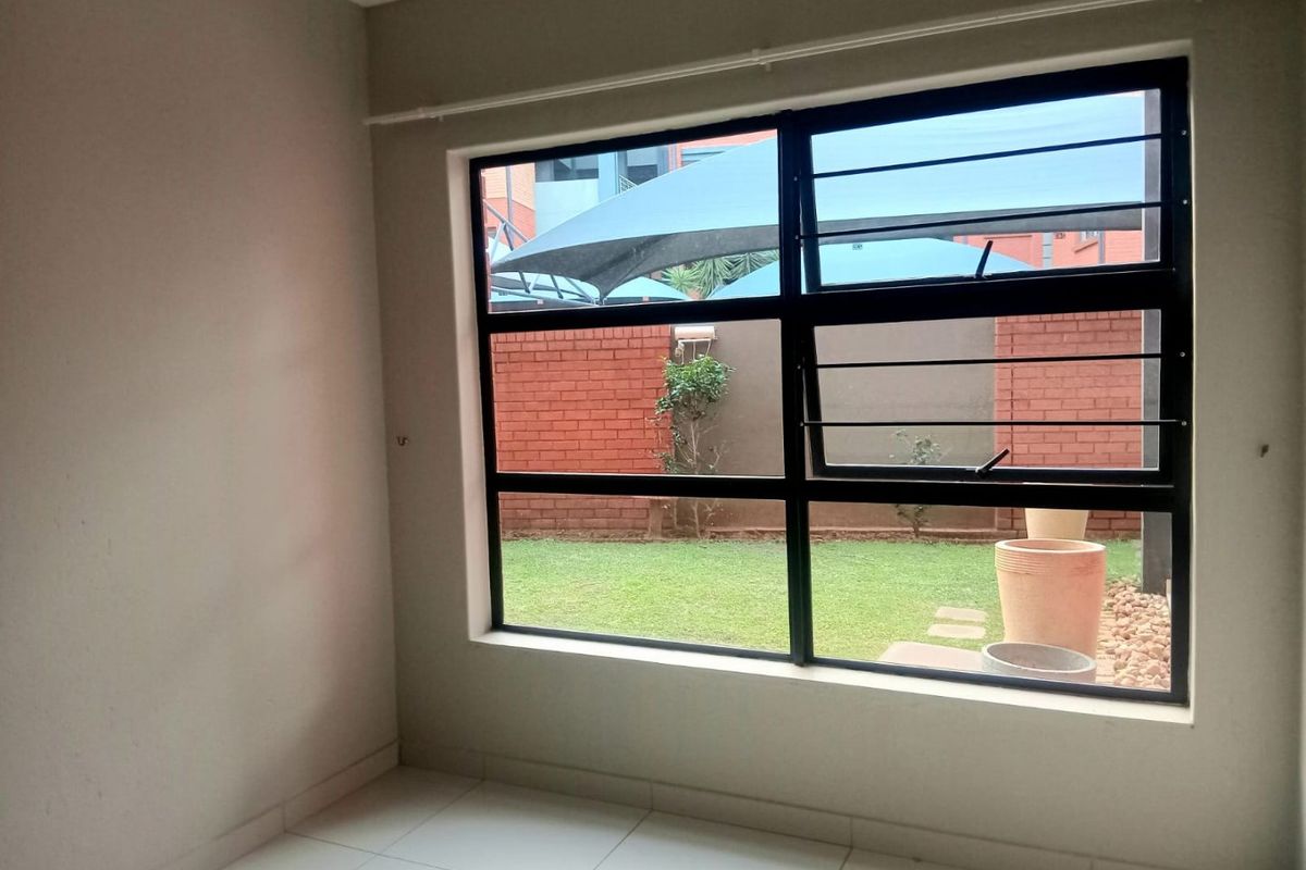 A bright, empty bedroom featuring tiled flooring, a large black aluminium window with security bars, and a view of the rear garden.