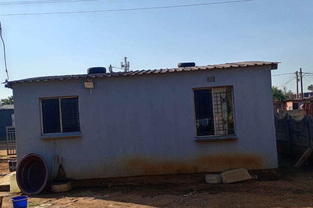 Rear exterior view of a single-story dwelling featuring a corrugated metal roof, security-barred windows, and visible exterior wall staining.