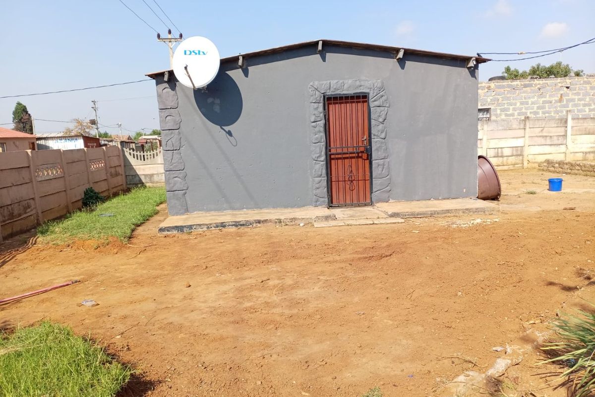 Rear view of a detached structure featuring a grey painted facade, security gate, and satellite dish, set within an unpaved yard with boundary walls.