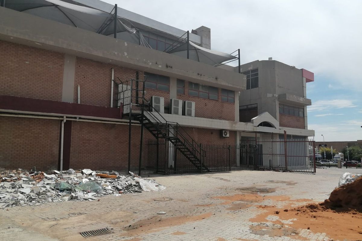 Rear exterior view of a commercial building featuring brick facade, metal staircase, security fencing, and a paved yard area with visible debris.