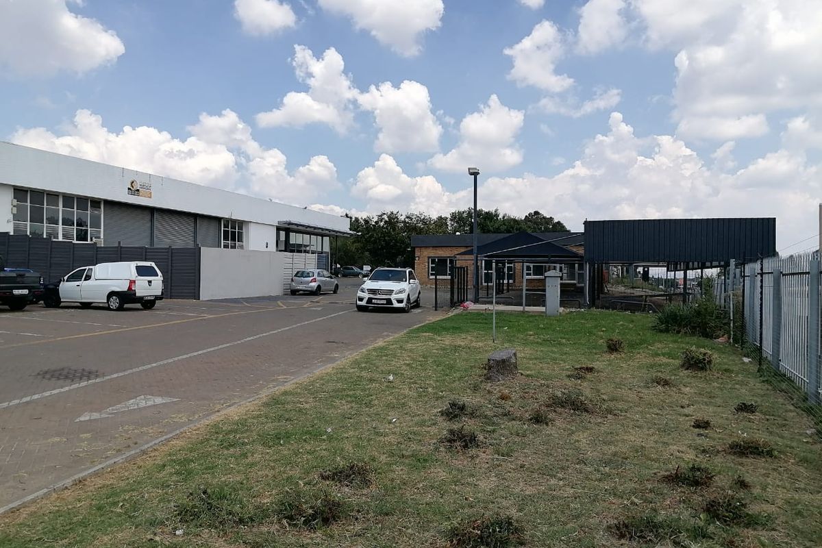 Exterior view of a commercial property featuring a paved parking area, perimeter fencing, and a landscaped lawn with a metal structure.