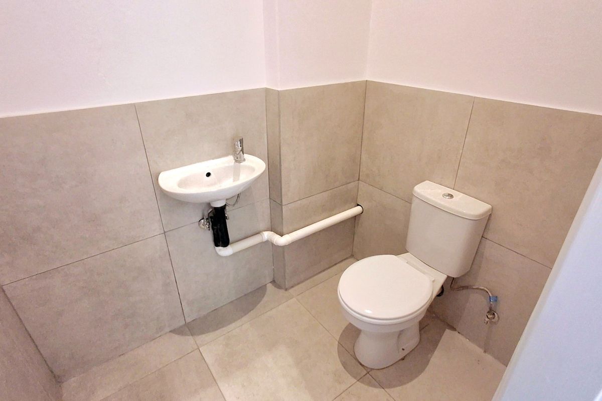 A compact guest bathroom featuring neutral ceramic tiling, a wall-mounted basin, and a standard toilet with exposed plumbing.