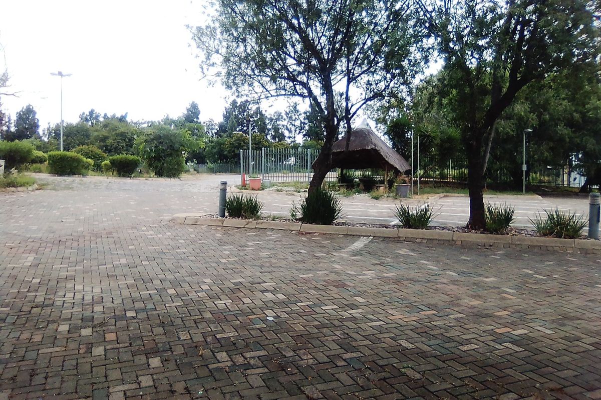 An outdoor area featuring a paved parking surface, mature trees, and a thatched gazebo structure within a fenced commercial property setting.