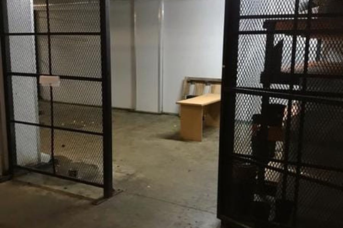 A utilitarian storage or cold room space featuring industrial metal mesh security gates, concrete flooring, and an overhead cooling unit.