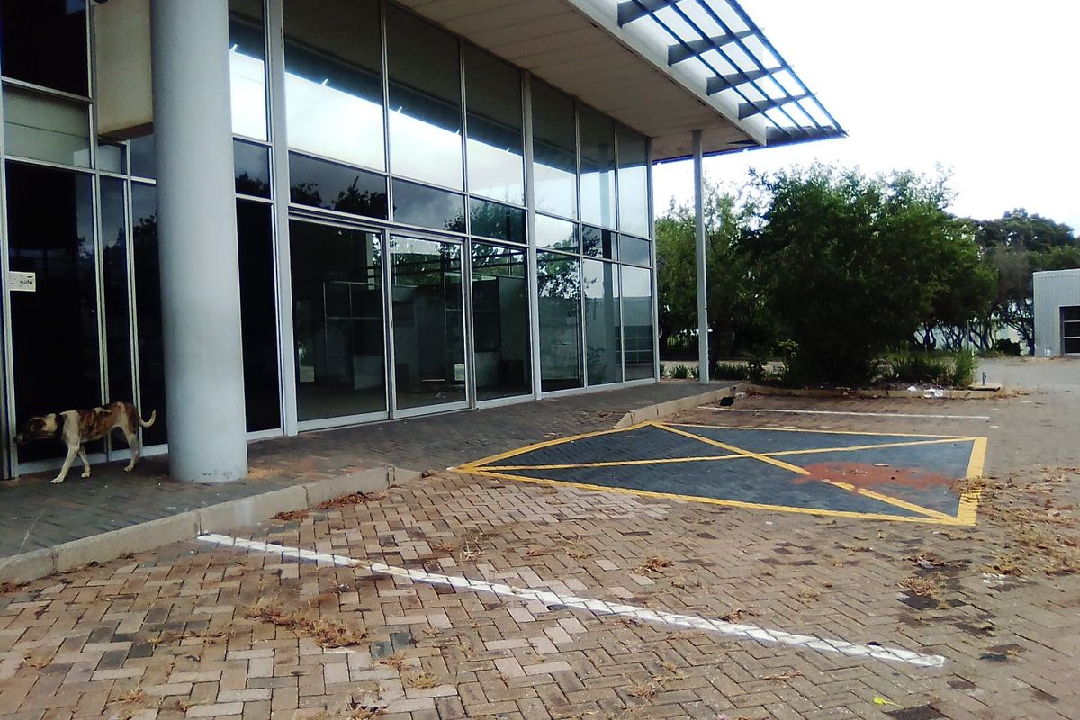 Front exterior view of a commercial building featuring a glass facade, structural columns, and a paved parking area.