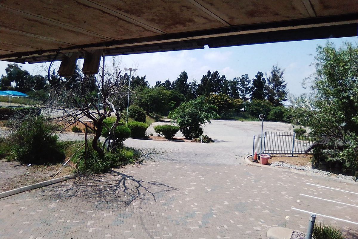 An outdoor view of a paved commercial driveway and parking area, featuring metal security fencing and a covered patio structure.