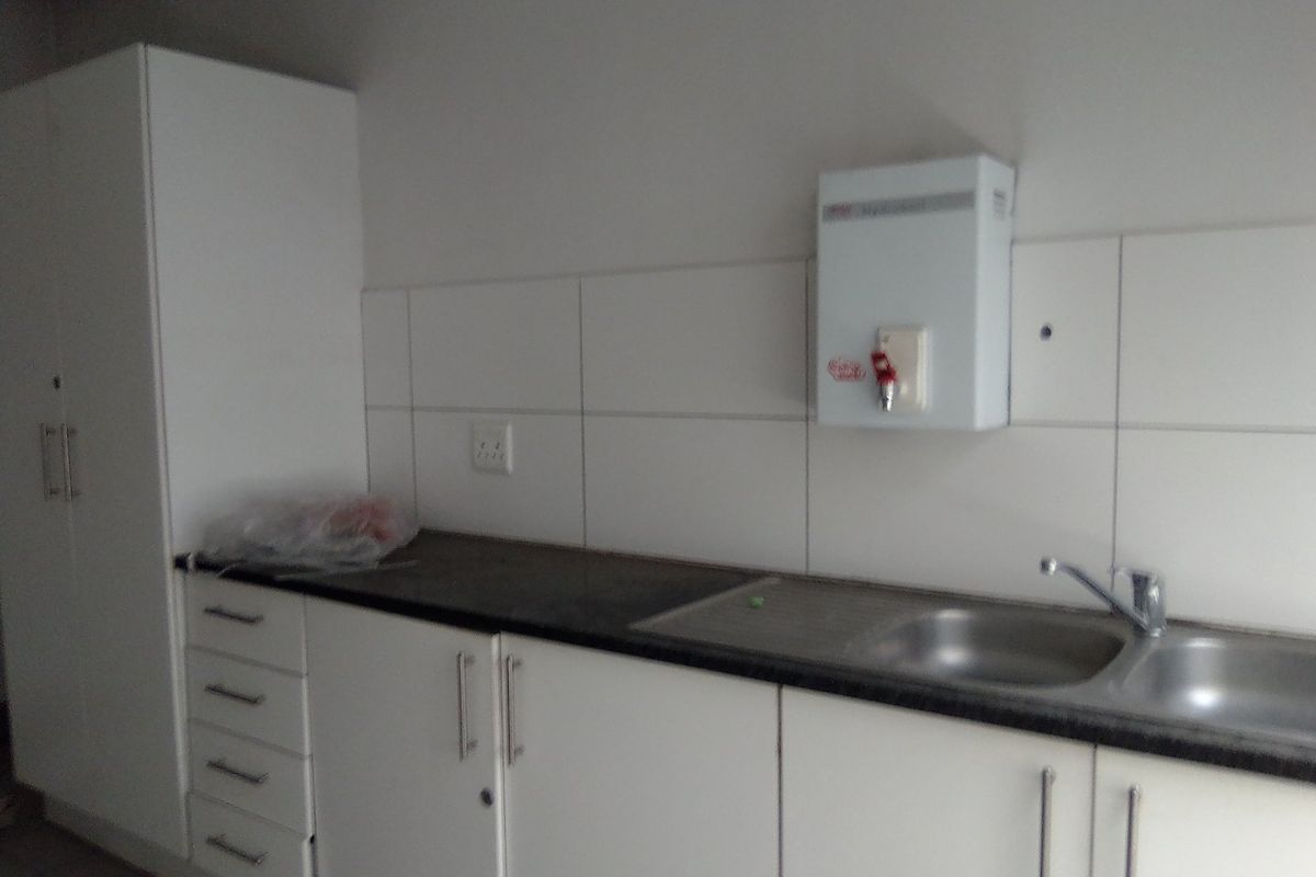 A functional kitchenette featuring white built-in cabinetry, a stainless steel sink, and a wall-mounted water heater with tiled backsplash.
