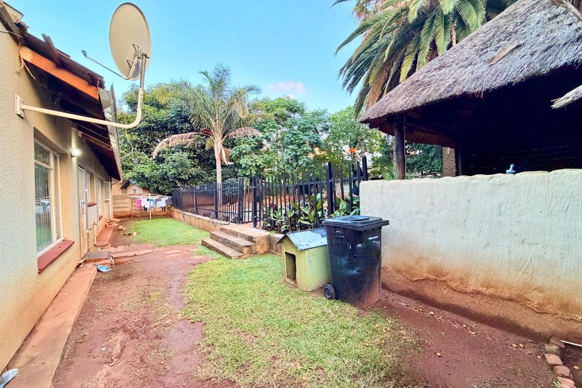 Rear garden area featuring a thatched lapa, metal perimeter fencing, and a side pathway along the main house structure.