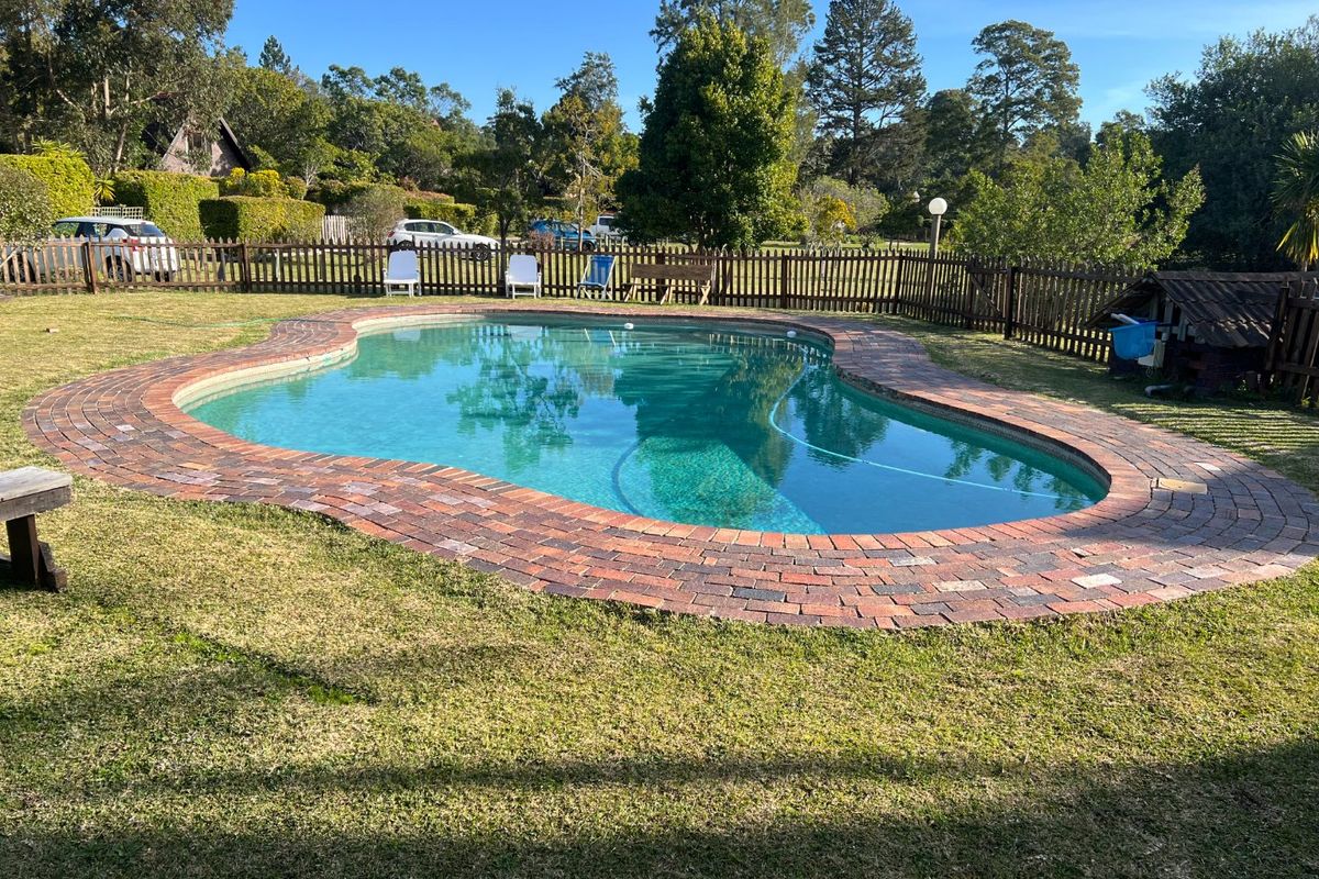 A well-maintained swimming pool featuring brick coping, surrounded by a lush garden and enclosed by a timber picket fence.