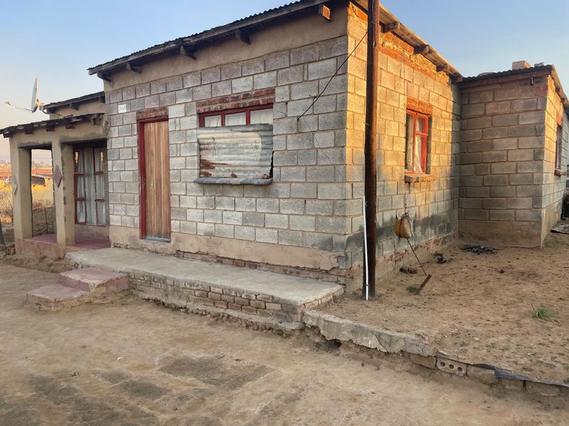 Front elevation of a single-story concrete block house featuring a small porch, corrugated iron roof, and basic metal window frames.