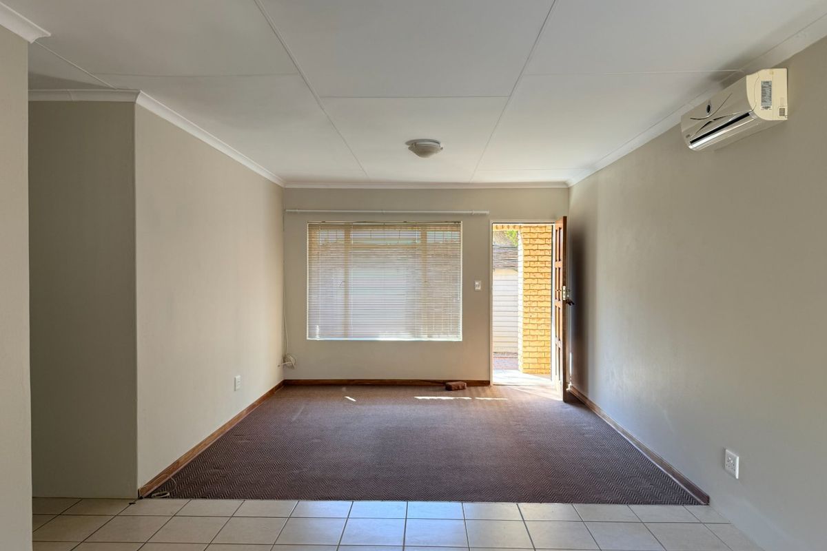 A spacious, empty living area featuring neutral walls, a wall-mounted air conditioning unit, tiled flooring transition to carpet, and direct access to the exterior.
