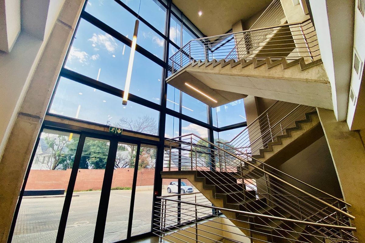 Modern commercial building entrance featuring a double-volume glass facade, concrete staircase with stainless steel railings, and polished flooring.