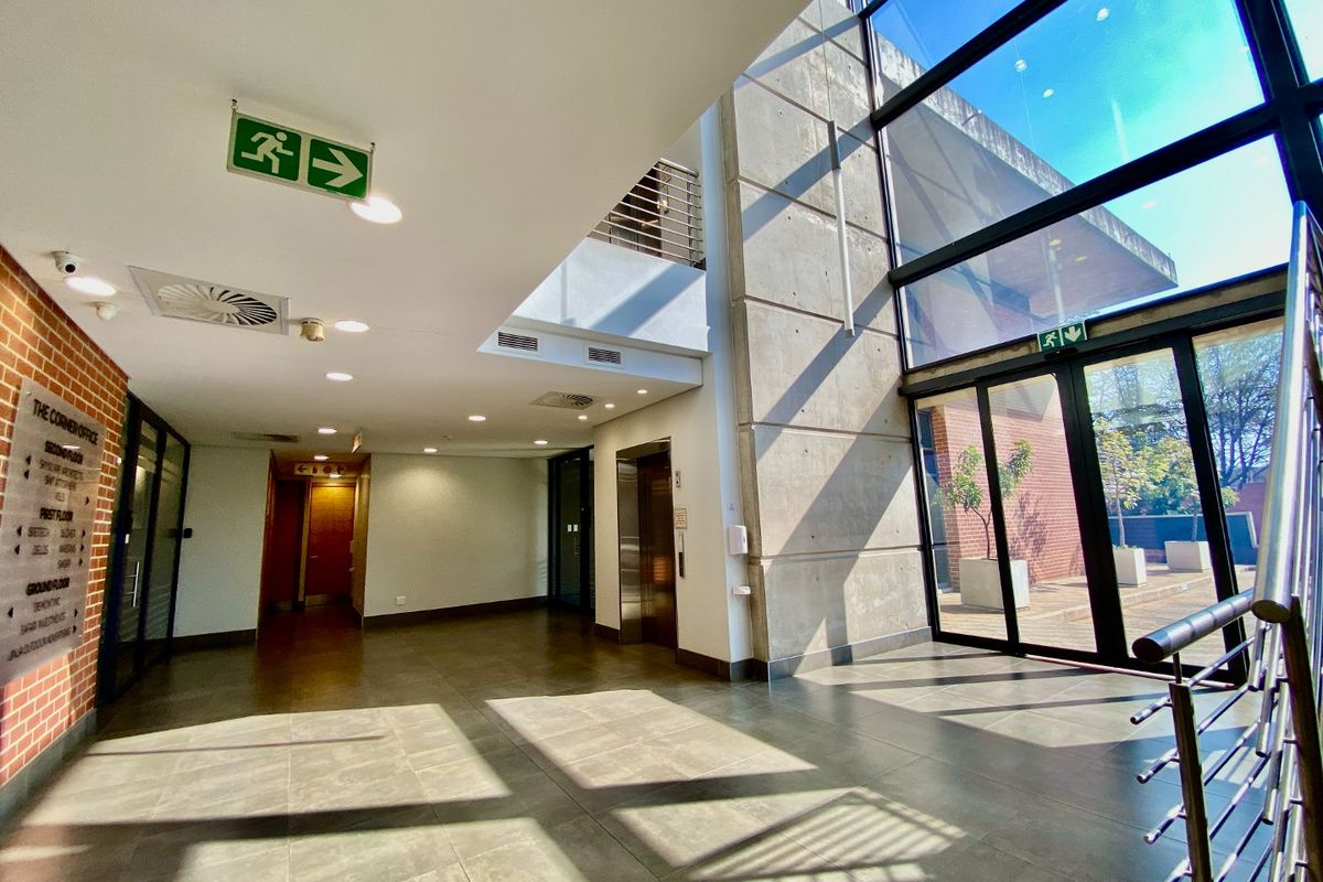 Modern commercial building lobby featuring double-volume glass facade, exposed concrete wall, tiled flooring, and elevator access.