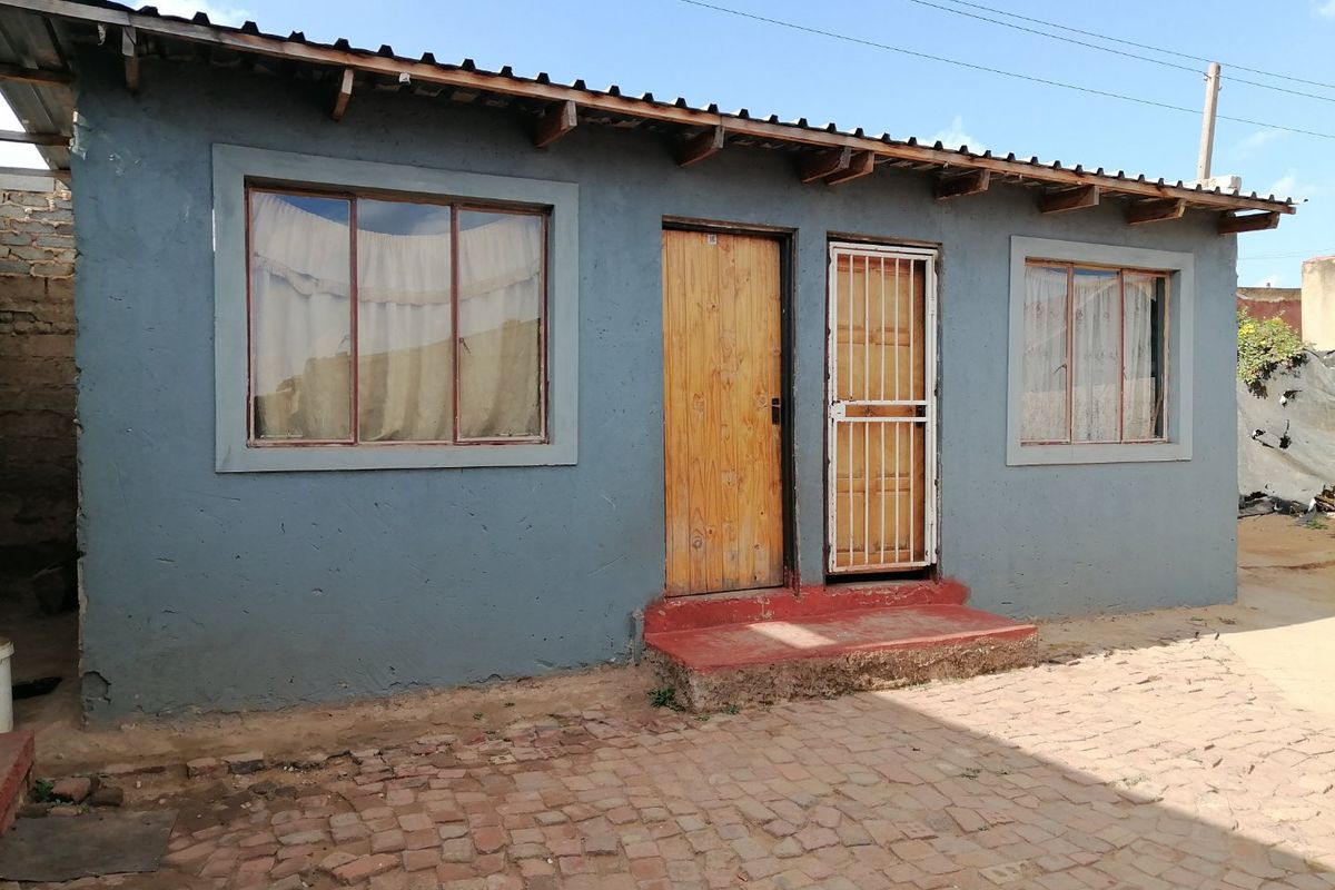 Exterior view of a single-story house featuring a painted facade, wooden door, security gate, and corrugated metal roof.