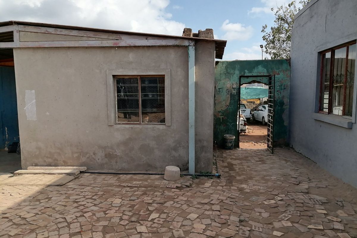 An exterior view of a residential property featuring a paved courtyard area, weathered exterior walls, and a gated access point to the rear.
