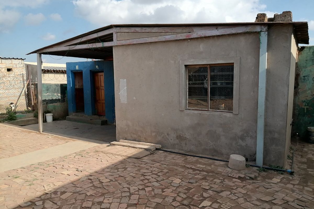 Exterior view of a modest dwelling featuring a covered patio area, concrete block construction, and paved courtyard space.