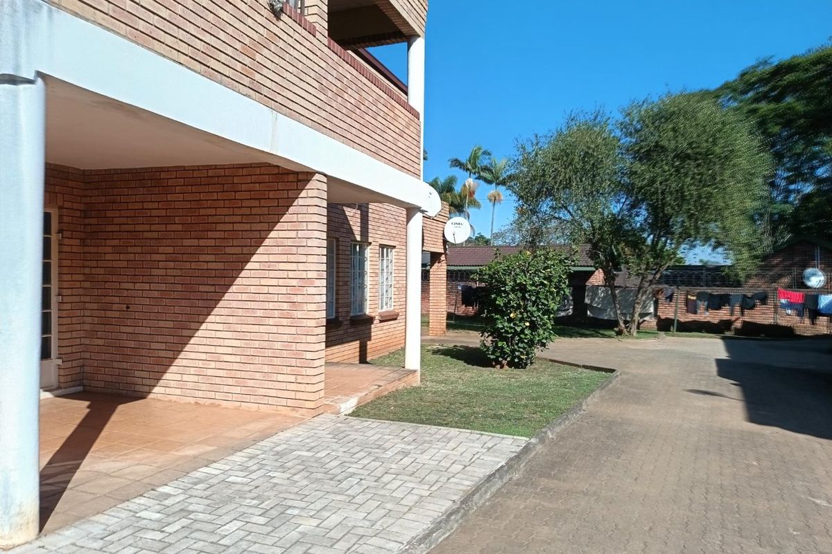 Exterior view of a brick apartment building featuring a covered patio area, paved walkway, and surrounding landscaped garden space.