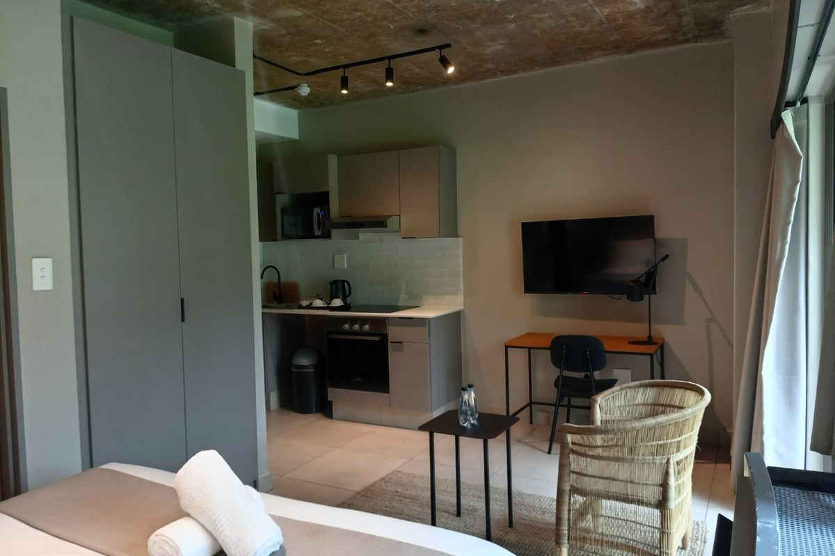 A modern open-plan studio apartment featuring a compact kitchenette with built-in oven, hob, and extractor, complemented by an exposed concrete ceiling.