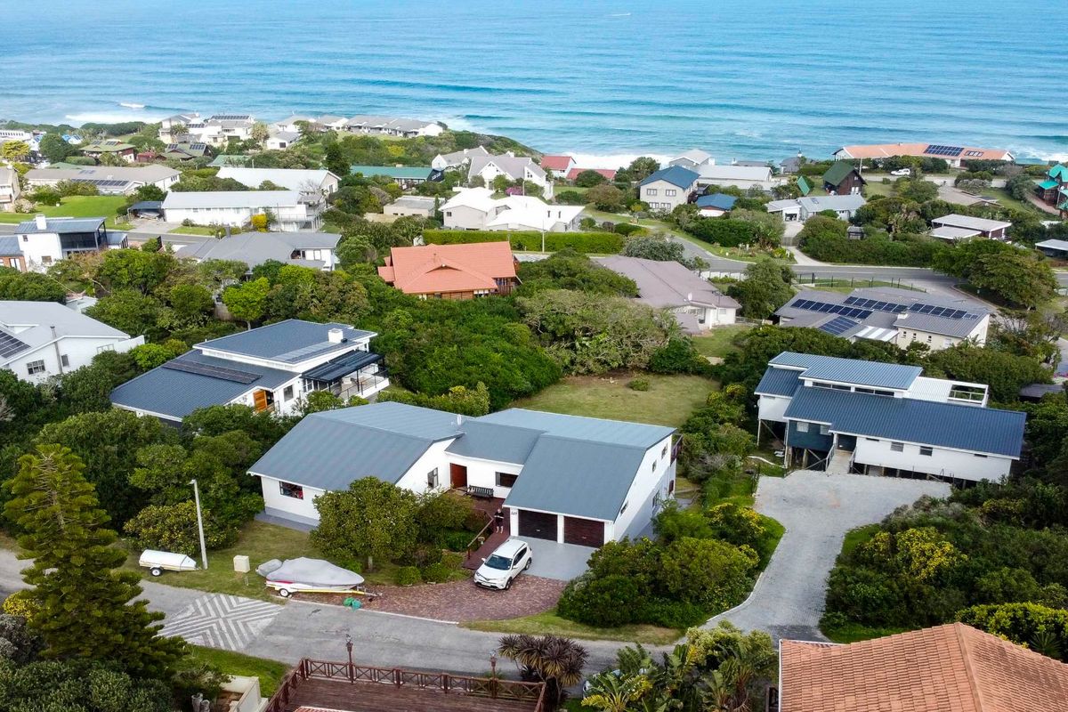 Aerial view of a coastal residential property featuring a modern home with a metal roof, paved driveway, and surrounding landscaped garden.