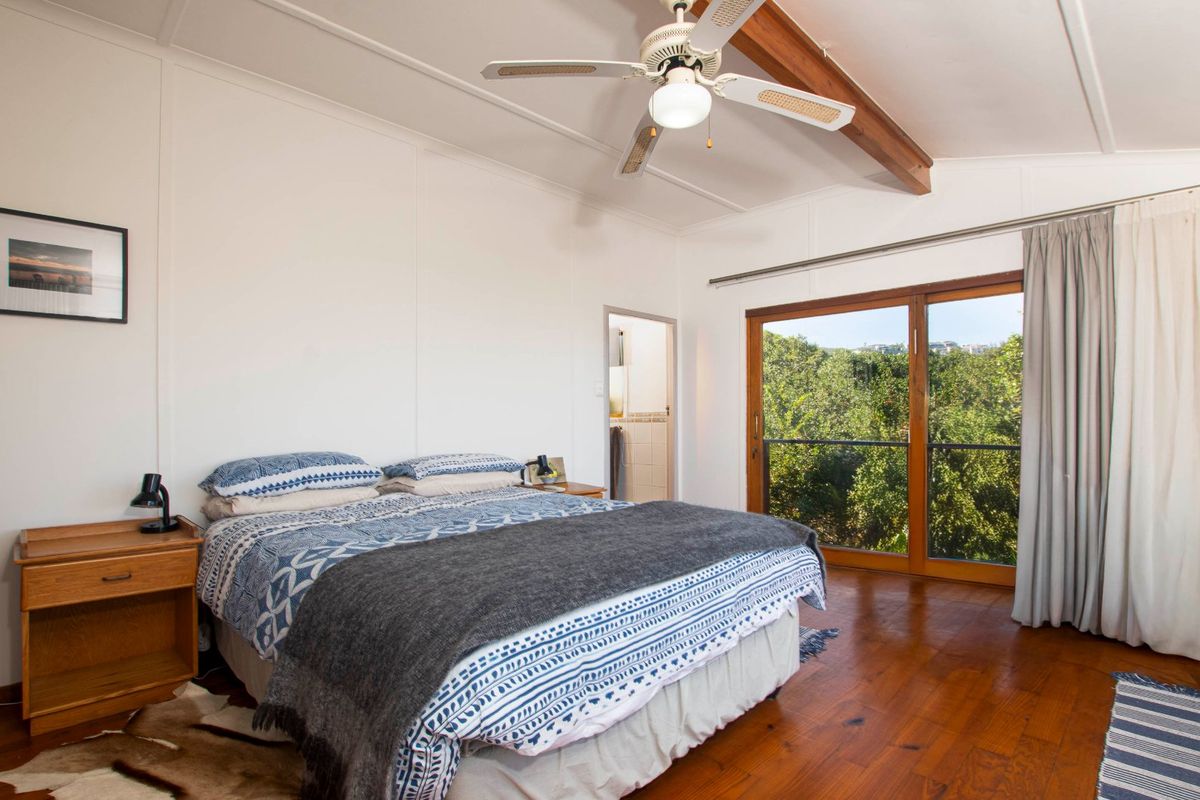Spacious bedroom featuring polished timber flooring, a vaulted ceiling with exposed timber beam, ceiling fan, and sliding glass doors opening to a leafy view.