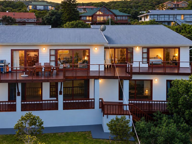 Rear elevation of a multi-level house featuring extensive timber decking, sliding glass doors, and a corrugated metal roof set against a lush hillside.