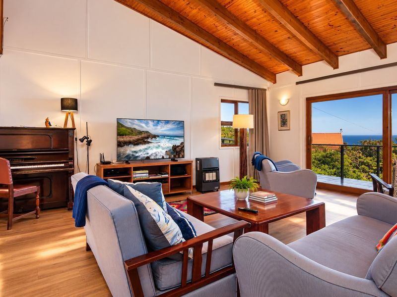 Spacious living room featuring a high vaulted timber ceiling, laminate flooring, and large sliding doors opening to a balcony with scenic coastal views.